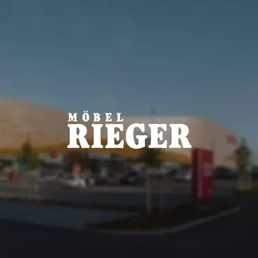 Auf dem Bild ist im Vordergrund das Logo von Möbel Rieger zu sehen und im Hintergrund ein Firmengebäude.